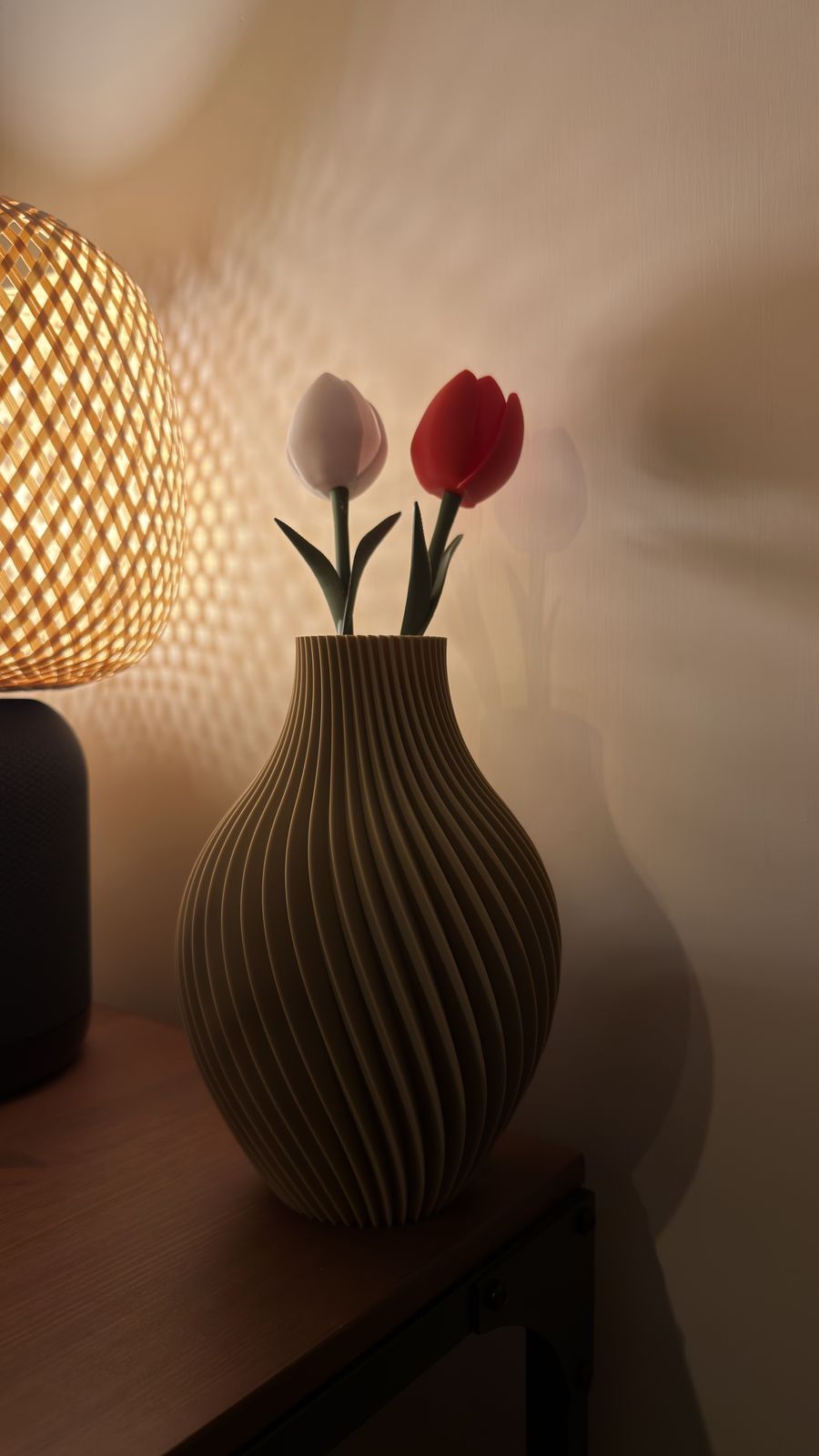 Due tulipani stampati in 3D, uno bianco e uno rosso, in un vaso a coste, illuminati di lato da una lampada di vimini